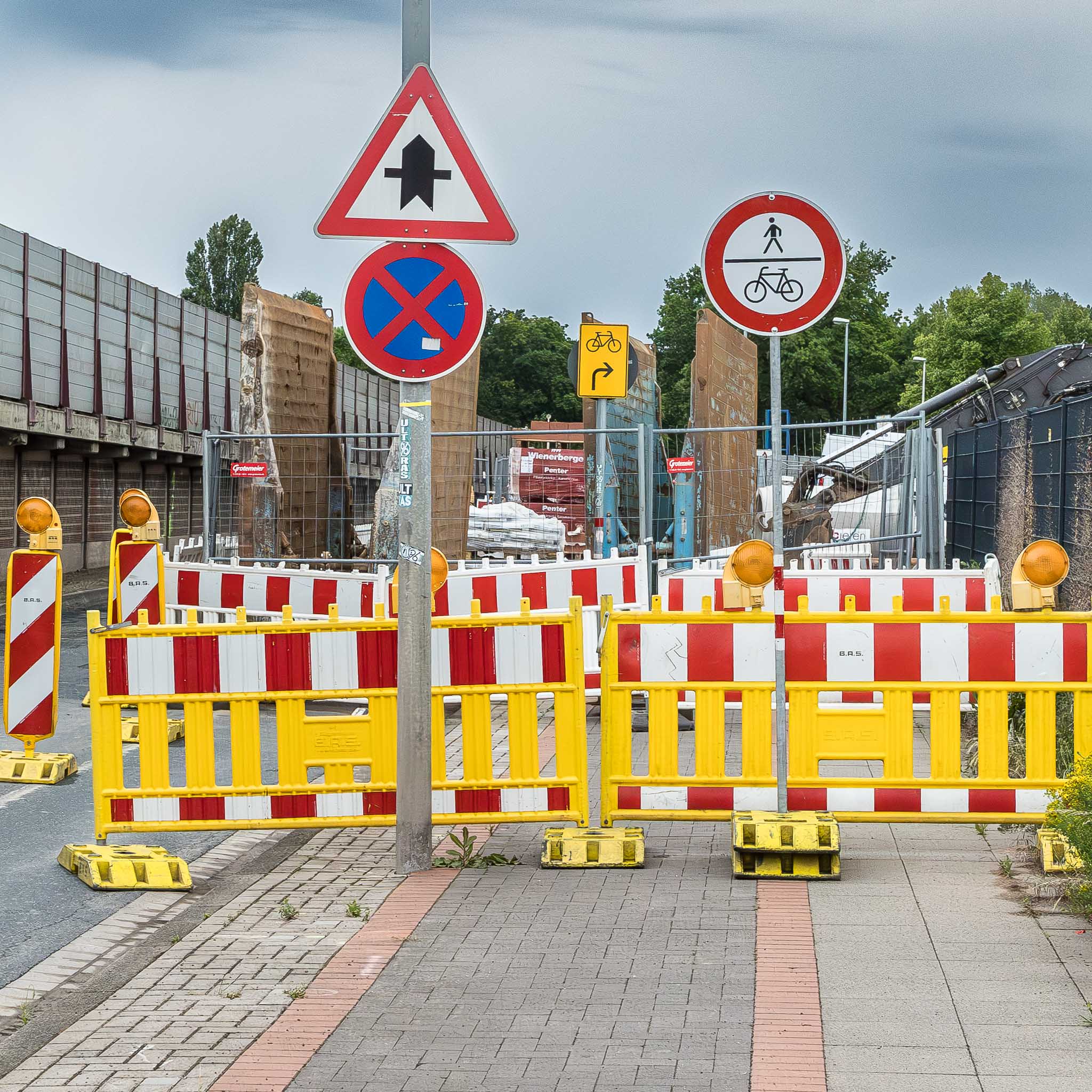 Baustellenabsperrung Zahlreiche rotweiße Baken und Absperrungen mit Blinkleuchten verhindern den Durchgang auf einem Fuß- und Radweg vor einer Baustellenabstellfläche. Die vier Verkehrzeichen »Vofahrt nur an dieser Kreuzung, Absolutes Halteverbot, Verbot für Fußgehende und Radfahrende, sowie Radwegumleirung rechts machen die Verwirrung komplett.